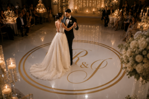 Bruidspaar danst op een luxe witte dansvloer met gouden initialen tijdens een romantische bruiloftsreceptie.